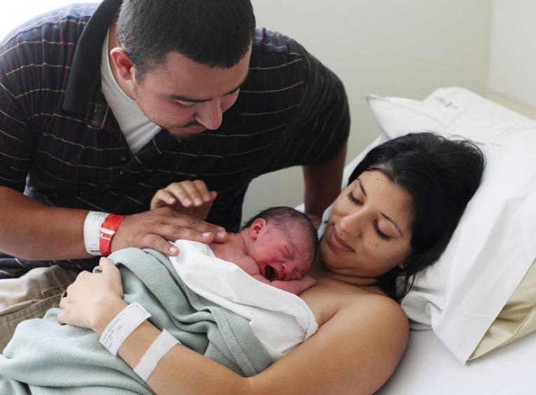 Mother lying in hospital bed holding newborn baby, with male partner watching over.