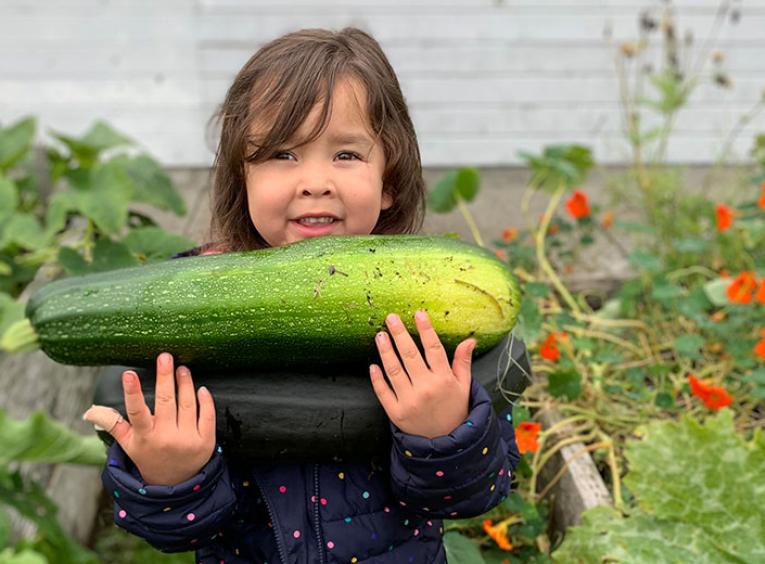 Little girl with dark hair in garden holding large zucchini