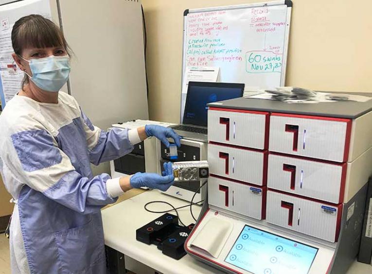 Woman in lab clothes and mask stands next to lab equipment