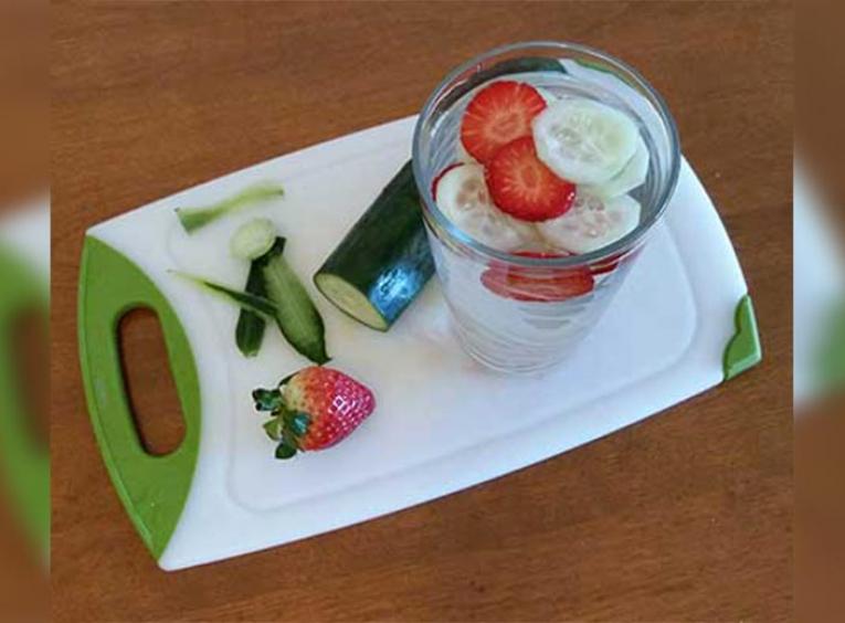 A cucumber and strawberry on a cutting board with slices in a glass of water.