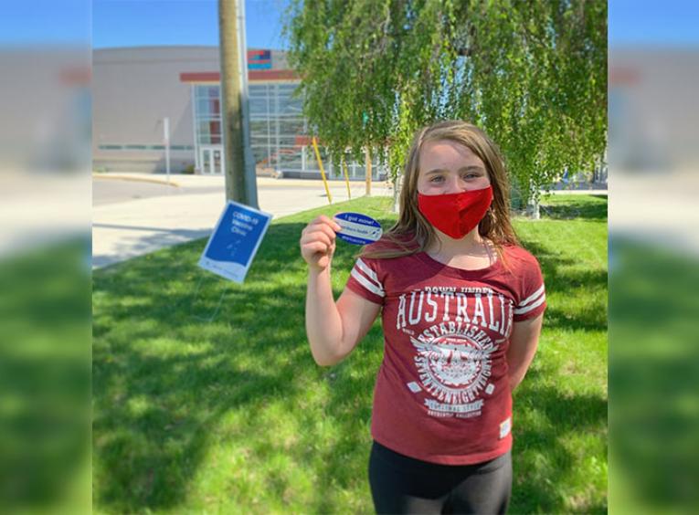 Young girl wearing a red mask while standing on the grass holding her COVID-19 vaccination sticker