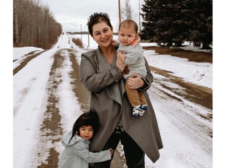 Woman holds baby on snowy country road while another child holds onto her leg