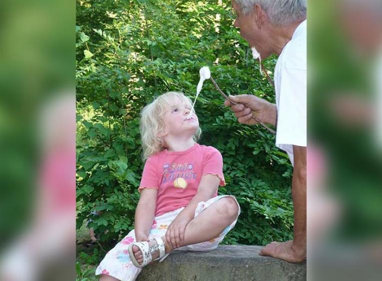 Child sitting on a log eating a marshmallow from her grandfather.