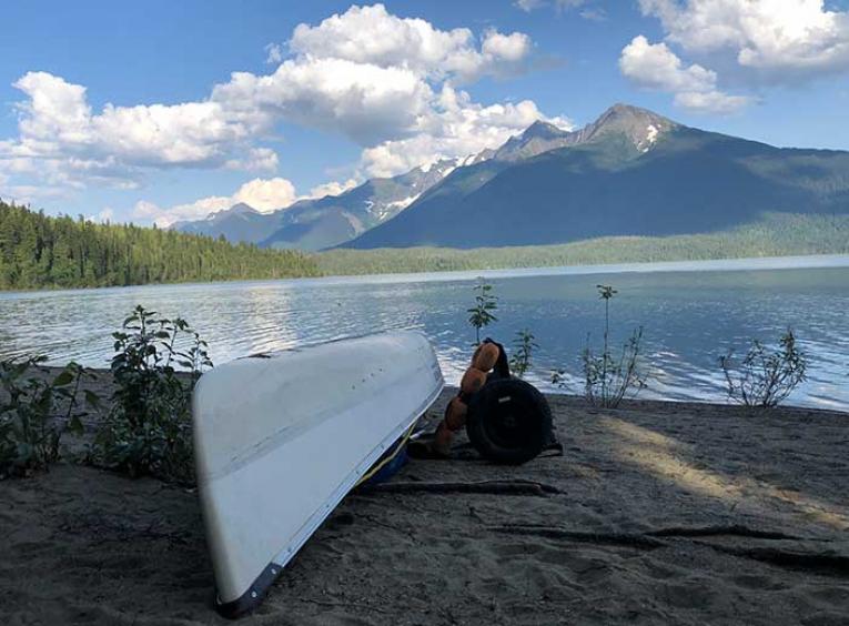 Lake view with a canoe resting on a sandy beach with mountains in the background.