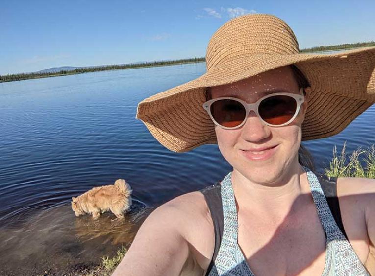 Woman with a hat and sunglasses stands by a lake with a dog. 