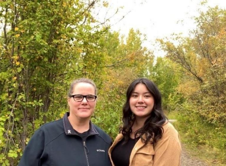 two women face the camera smiling there is a fall forest in the background