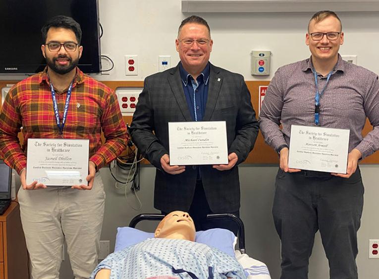 Three men stand in a row presenting their certificates over a medical dummy