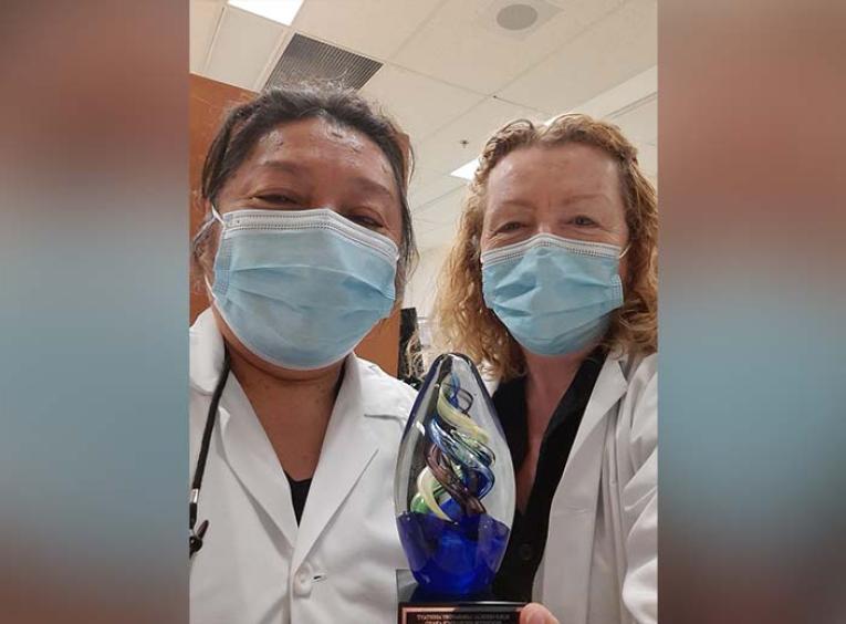 two women in medical masks and white lab coats hold up the excellence award