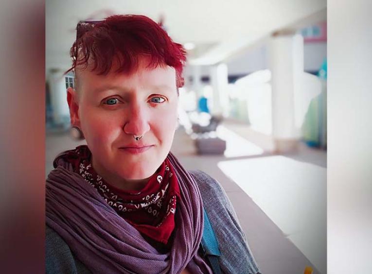 a person with short dyed-red hair smiles into the camera. They are wearing a cozy red scarf.