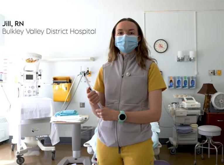 A picture of Jill - she is wearing yellow scrubs with a grey fleece vest on top. She has long light brown hair and is wearing a blue surgical mask.