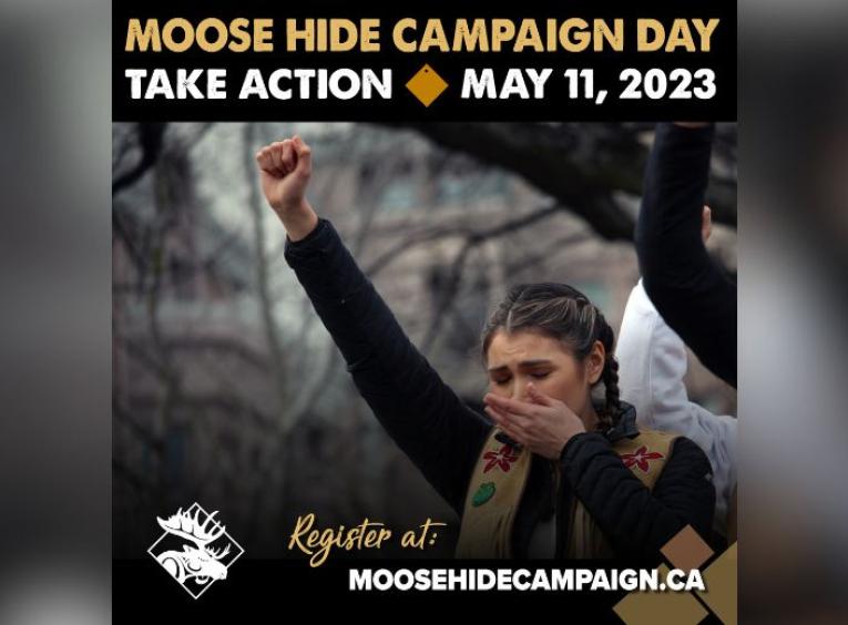 Moose hide campaign supporter standing with one hand over her mouth while the other is a raised fist in the air at a campaign event
