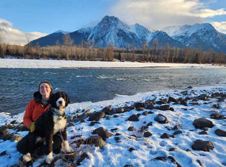 Woman and her dog in winter setting, beside a river with mountains in the background