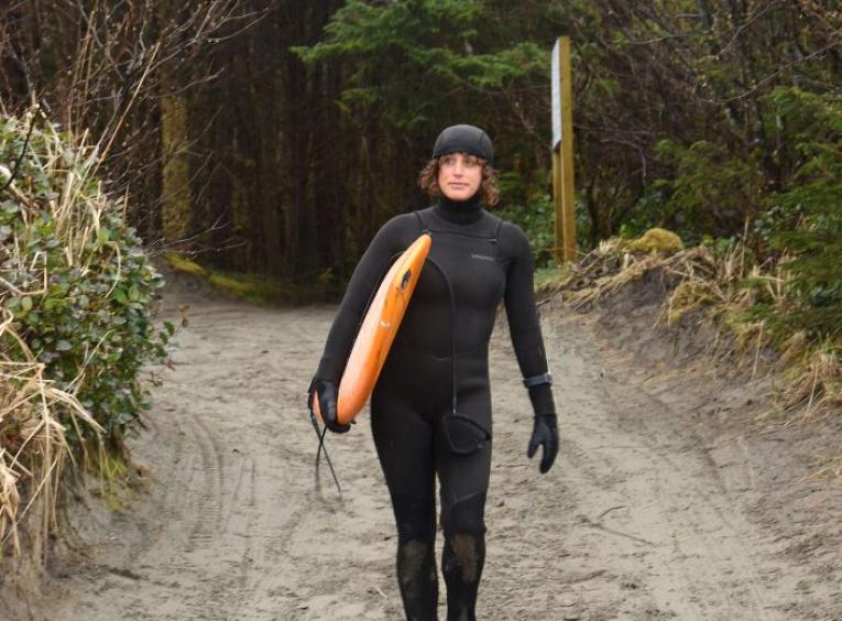 Dr. Jocelyn Black, head to toe in a black wet suit, walking down a beach pathway toward the water, holding her surf bard under her right arm.