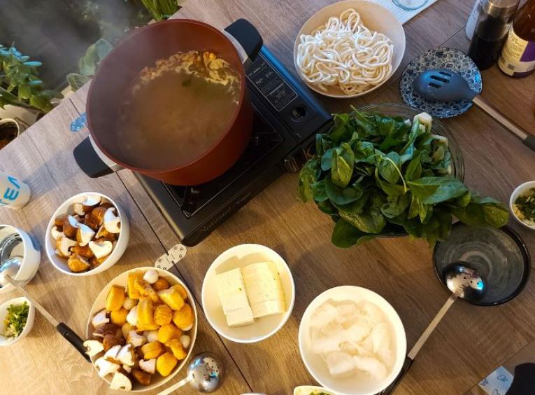 Several bowls containing noodles, tofu, greens, sauces and a large pot sitting on a table