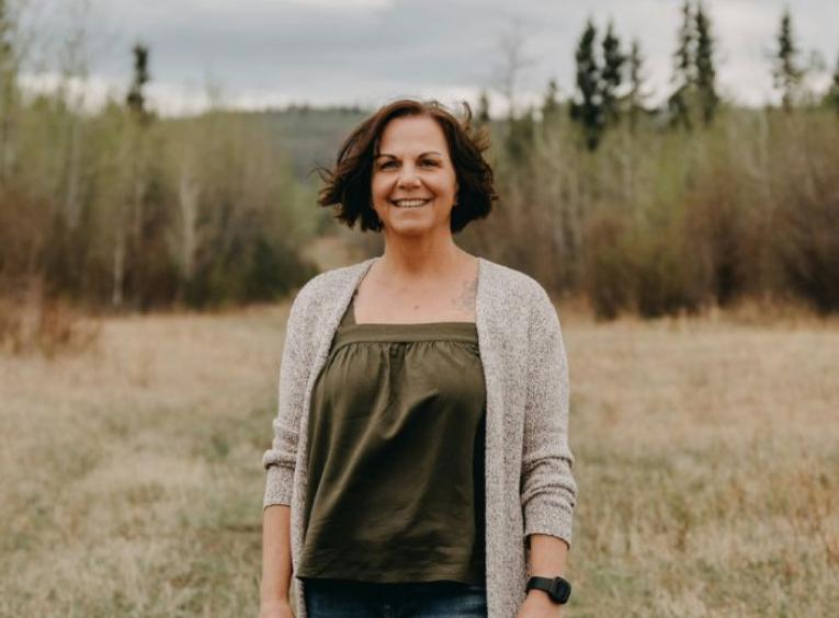 A woman in a green shirt with tan cardigan stands in a field in front of evergreen tress. 