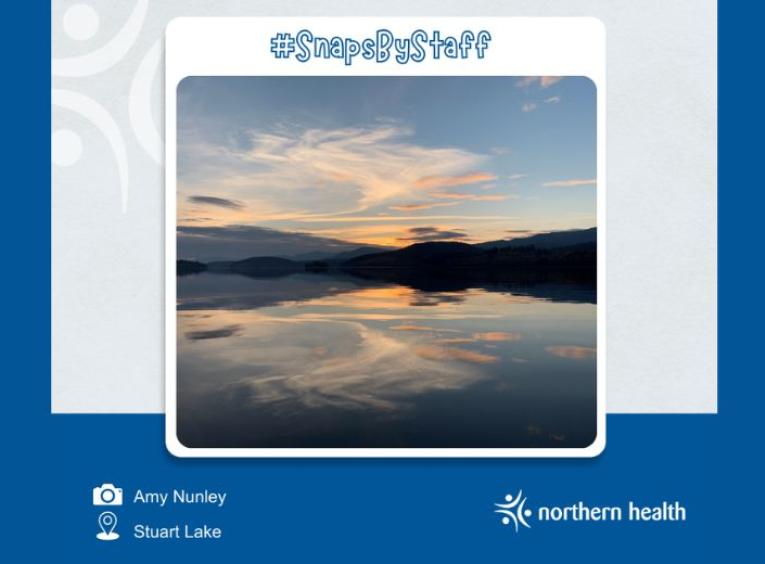 Calm waters reflecting clouds, shadows of mountains, and the orange setting sun of the background. 