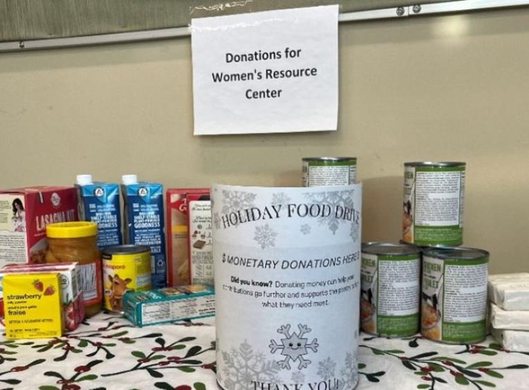 A stack of non-perishable food cans sits on a table covered with a holiday theme table cloth