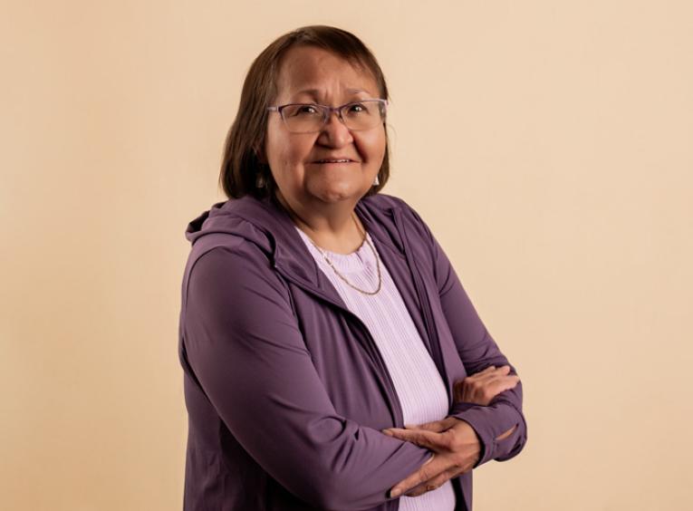 Indigenous woman stands with arms crossed across her chest and welcoming smile.