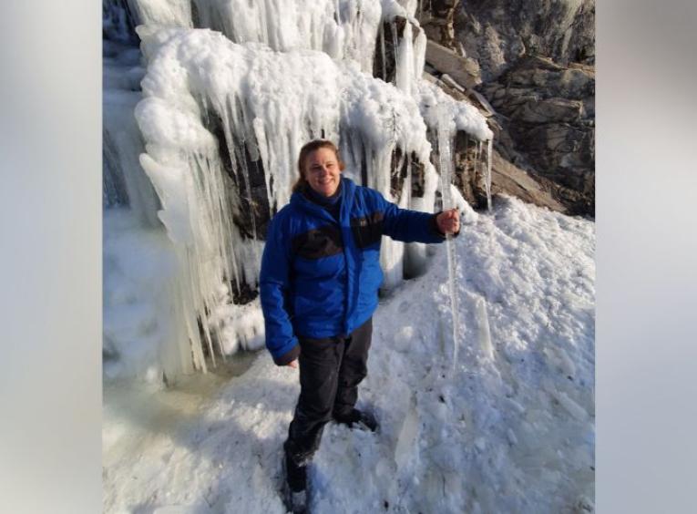 Penelope standing under a frozen waterfall wearing blue ice climbing gear