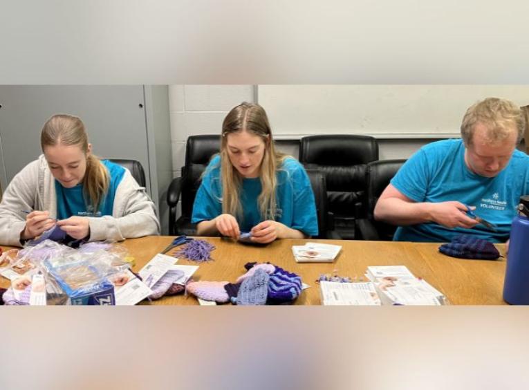 Volunteers knitting purple hats with supplies scattered across the table.