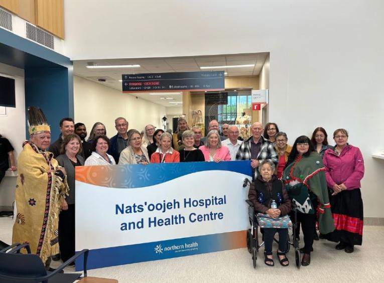 A large group of people stand holding a sign noting the name of the new hospital. 