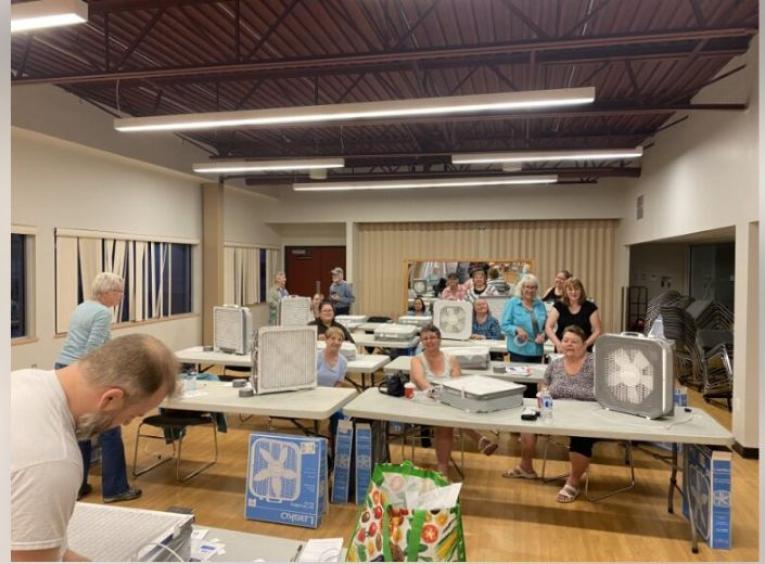 Participants sit at long tables with partially constructed DIY air cleaners standing next to them.
