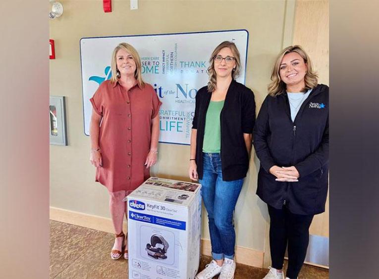 Three women stand beside a car seat box in a hallway in front of the Spirit of the North Healthcare Foundation sign.
