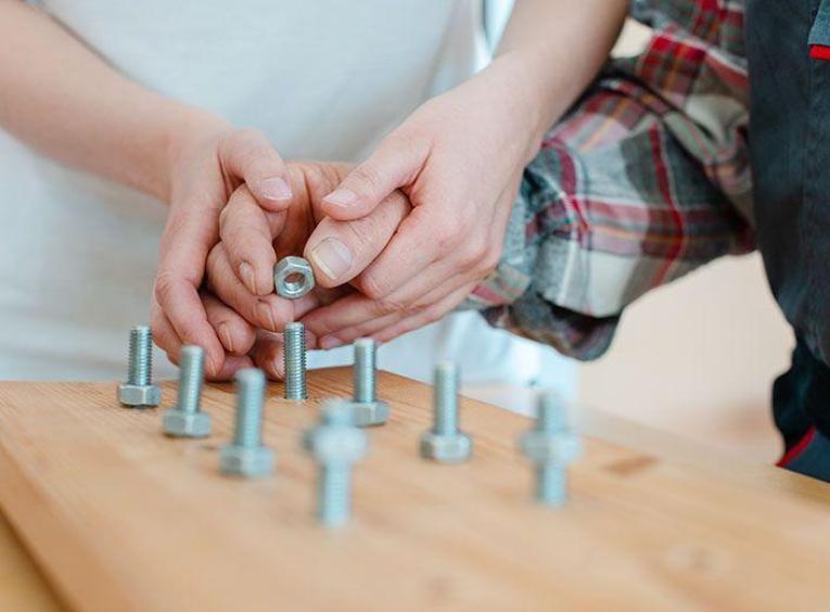 Two hands over a woodedn board attempting to screw nuts onto protruding bolts.