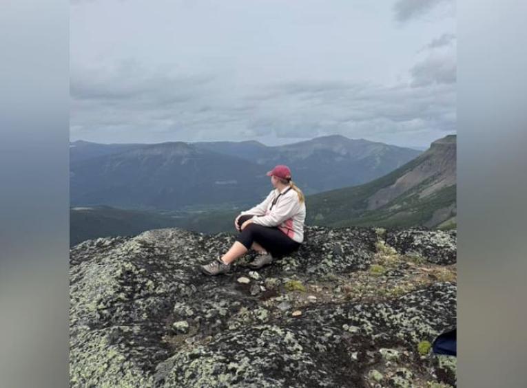 Amanda sits atop a mountain, viewing forests and mountains in the background. 