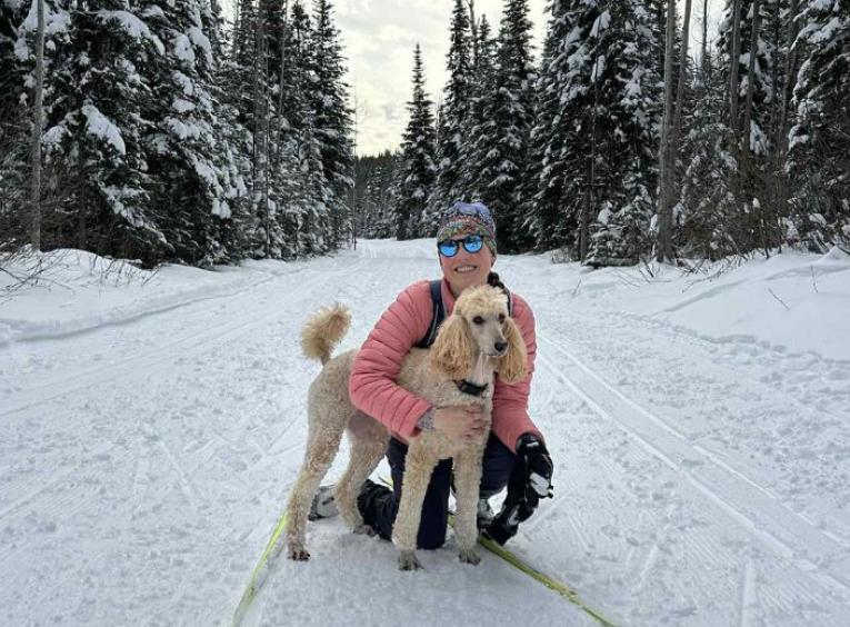 Haylee holding her dog on one knee in the snow, surrounded by trees