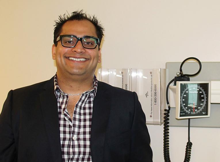 Dr. Abdulla, wearing a jacket and checked shirt, smiles in front of medical equipment. 