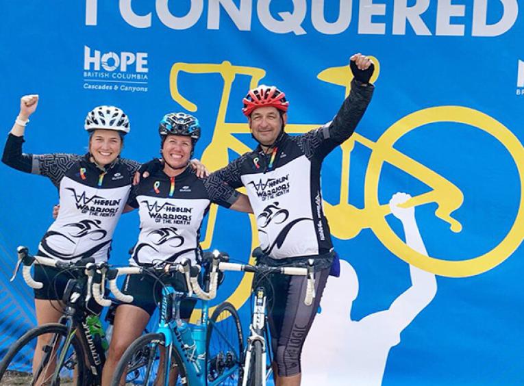 Haylee, her mom, and dad celebrate finishing the 2019 Ride to Conquer Cancer in their bike outfits with helmets on, and arms raised. 
