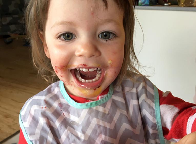 An adorable child, with food all over it's face, smiles into the camera and holds a peanut butter and jam sandwich. 