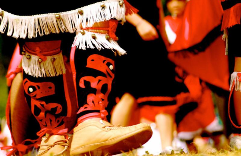 A person's legs are in the foreground, wearing moccasins, and traditional leggings. More similarly dressed people are blurred in the background.