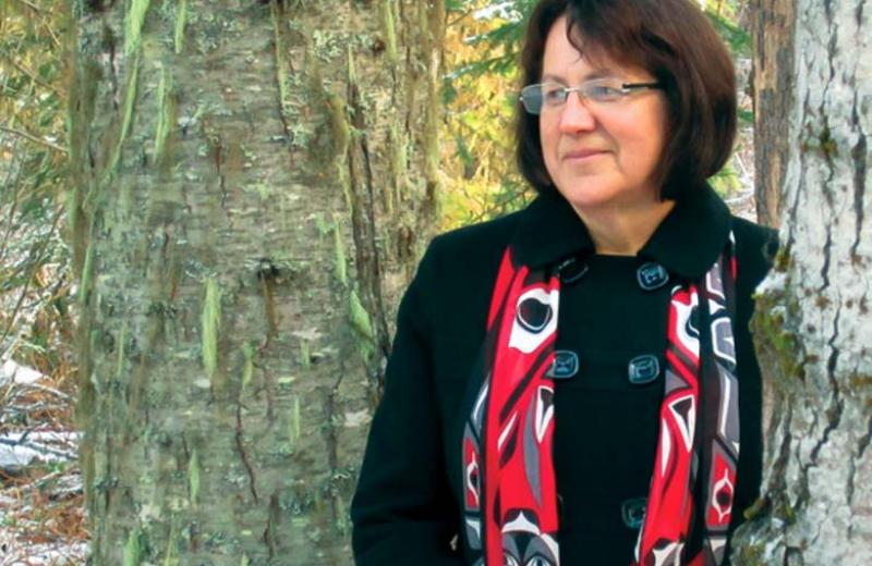 Dr. Margo Greenwood stands between two trees, wearing a scarf with Indigenous art on it.