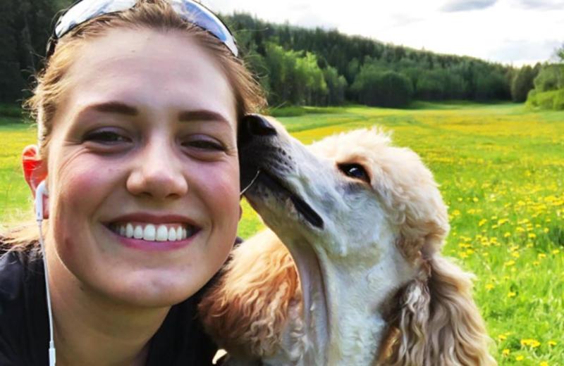 Haylee smiles for the camera as her dog sniffs the side of her head. A meadow is in the background.