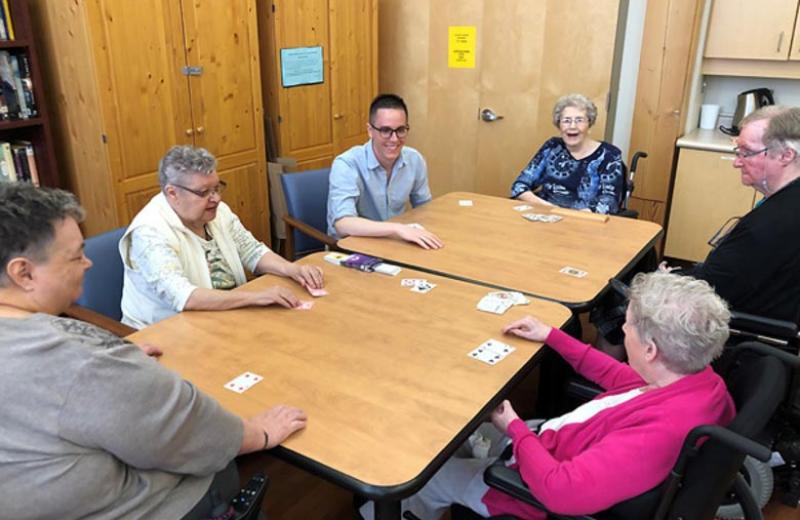Zach, a young man, plays cards with five seniors.