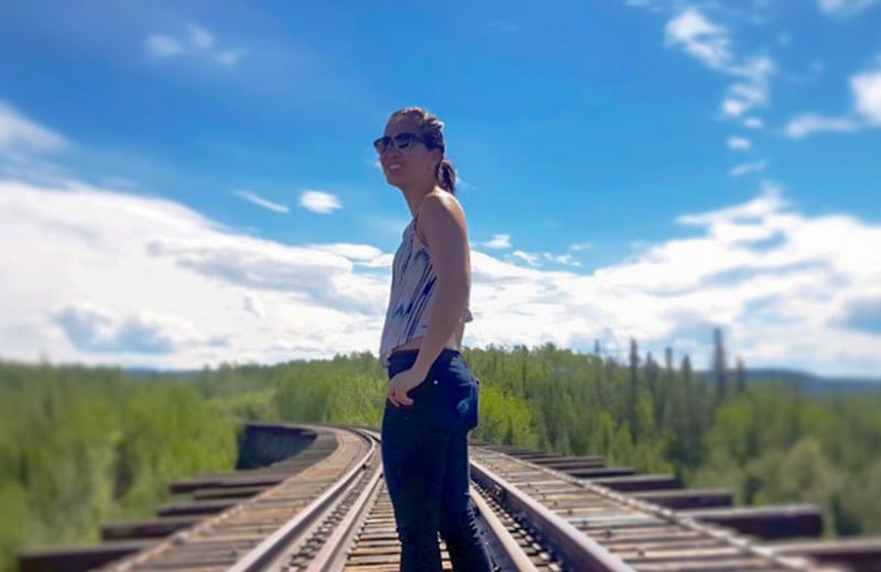 Melinda stands on a train track that disappears in the distant forest. A sunny sky beats down on her.