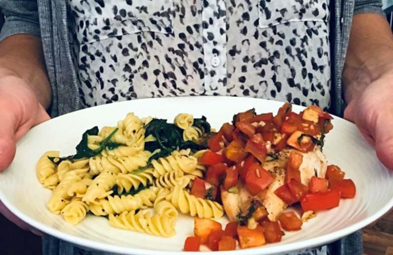 A person holds a white plate of food. On the left of the plate is pasta noodles with spinach, on the right is a chicken breast covered in chunks of tomato.