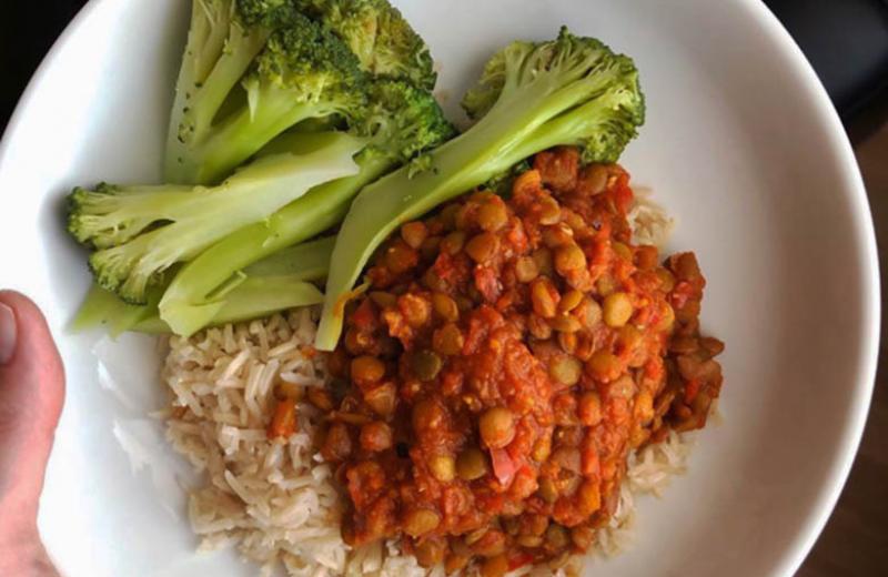 A hand holds a plate of Moroccan lentils on rice and broccoli.