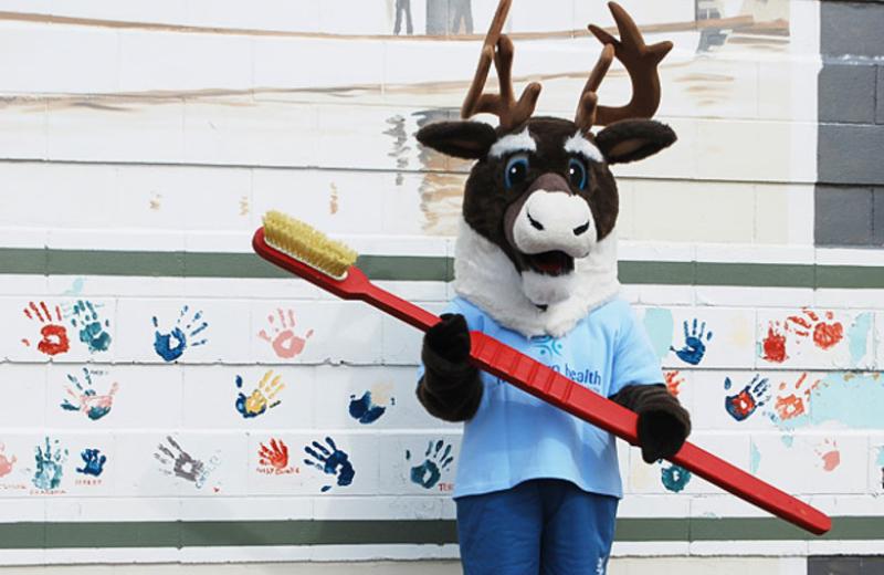 Spirit the Cariboo holding a large toothbrush, standing against a mural of handprints.