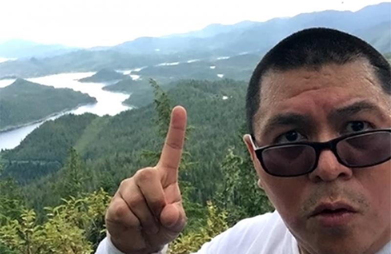 Man with index finger in the air with river and mountains in the background.