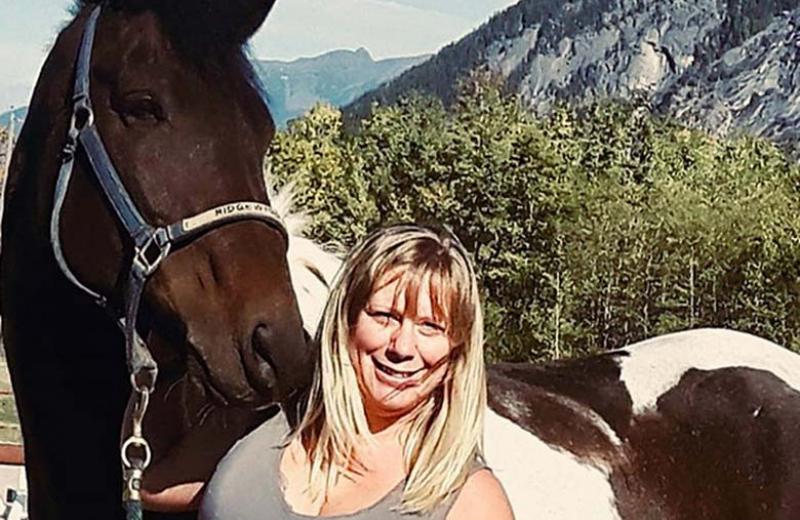Clare smiles into the camera. Her brown and white horse is directly behind her. Mountains and forest is further in the background.