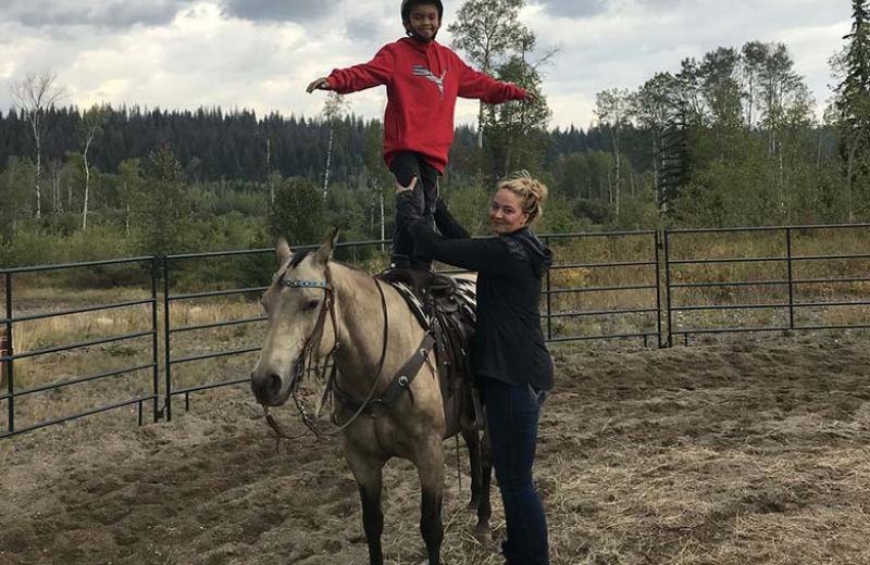 A woman poses, holding the legs of a young boy who is standing on top of a saddled horse in a field.