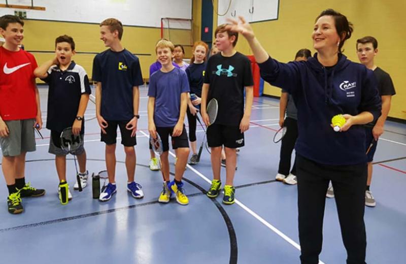 Woman tosses a badminton birdie in a gymnasium as a group of young students watches.