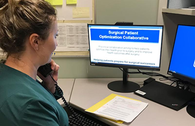 A side angle shot of a woman in a green sweater sitting at a desk in front of a computer screen that says "Surgical Patient Optimization Collaborative."