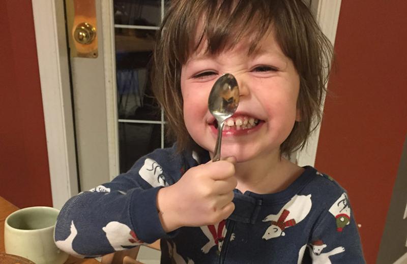 A young, smiling boy wearing polar bear pyjamas and sitting at a kitchen table holds a spoon up to his nose.