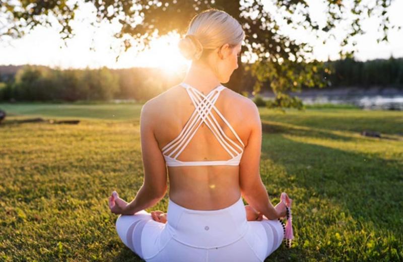 Woman practising yoga, sitting cross-legged on grass with her back to the camera. The sun sets in the horizon, over her left shoulder.