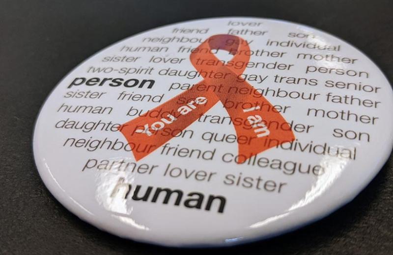 A World Aids Day button is pictured. The button features a red ribbon on a white background with words that humanize people battling HIV.