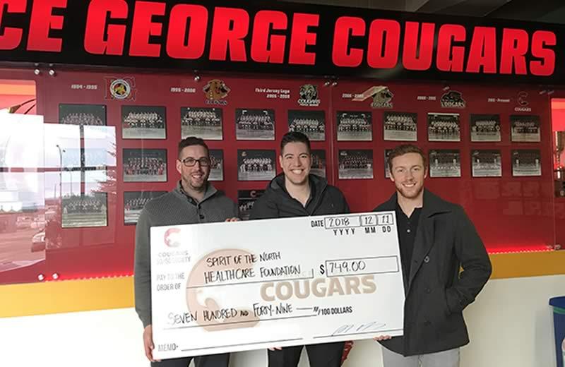 Three men holding a large cheque in front of a red sign Prince George Cougars.